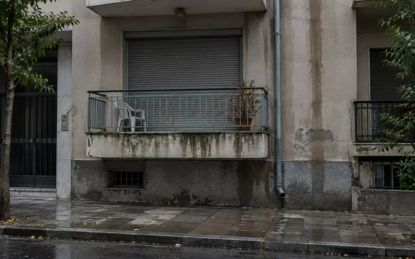 A first-floor Athens apartment with a closed roller shutter and a balcony chair after rain (illustration)