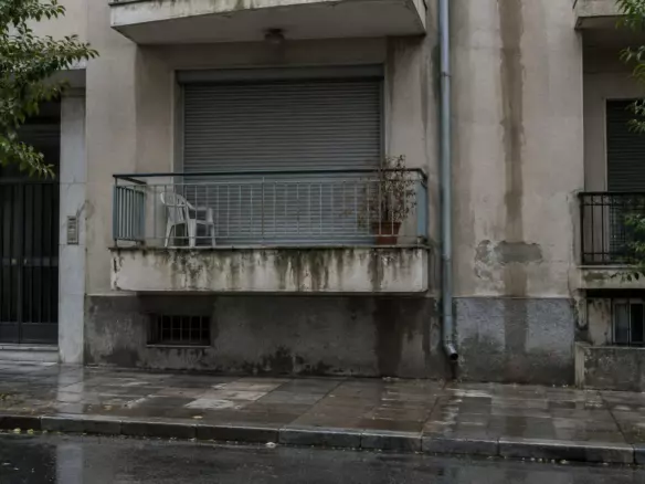 A first-floor Athens apartment with a closed roller shutter and a balcony chair after rain (illustration)