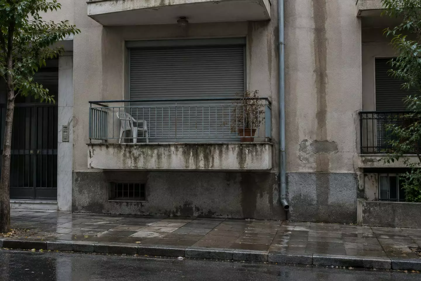 A first-floor Athens apartment with a closed roller shutter and a balcony chair after rain (illustration)