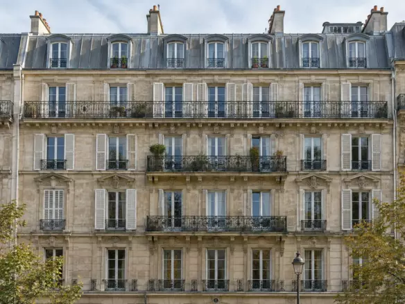 The facade of a Haussmann-era apartment building with wrought-iron balconies and tall shuttered windows on a Paris street in early autumn (illustration)