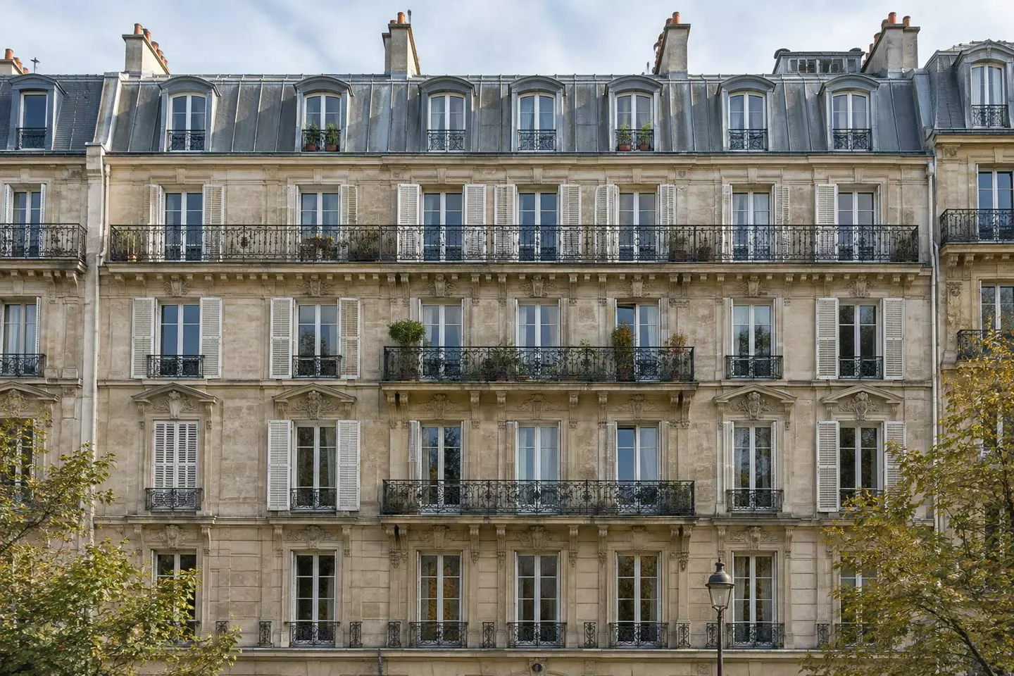 The facade of a Haussmann-era apartment building with wrought-iron balconies and tall shuttered windows on a Paris street in early autumn (illustration)