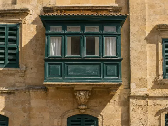 A traditional Maltese limestone balcony with a painted wooden box and shutters on a townhouse facade in early autumn (illustration)