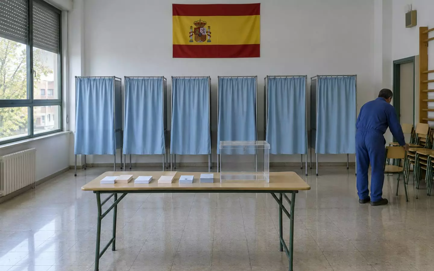 A polling station with blue-curtained booths and a transparent ballot box being prepared in a Madrid school hall in early autumn (illustration)