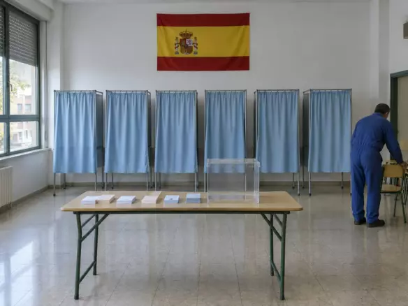 A polling station with blue-curtained booths and a transparent ballot box being prepared in a Madrid school hall in early autumn (illustration)