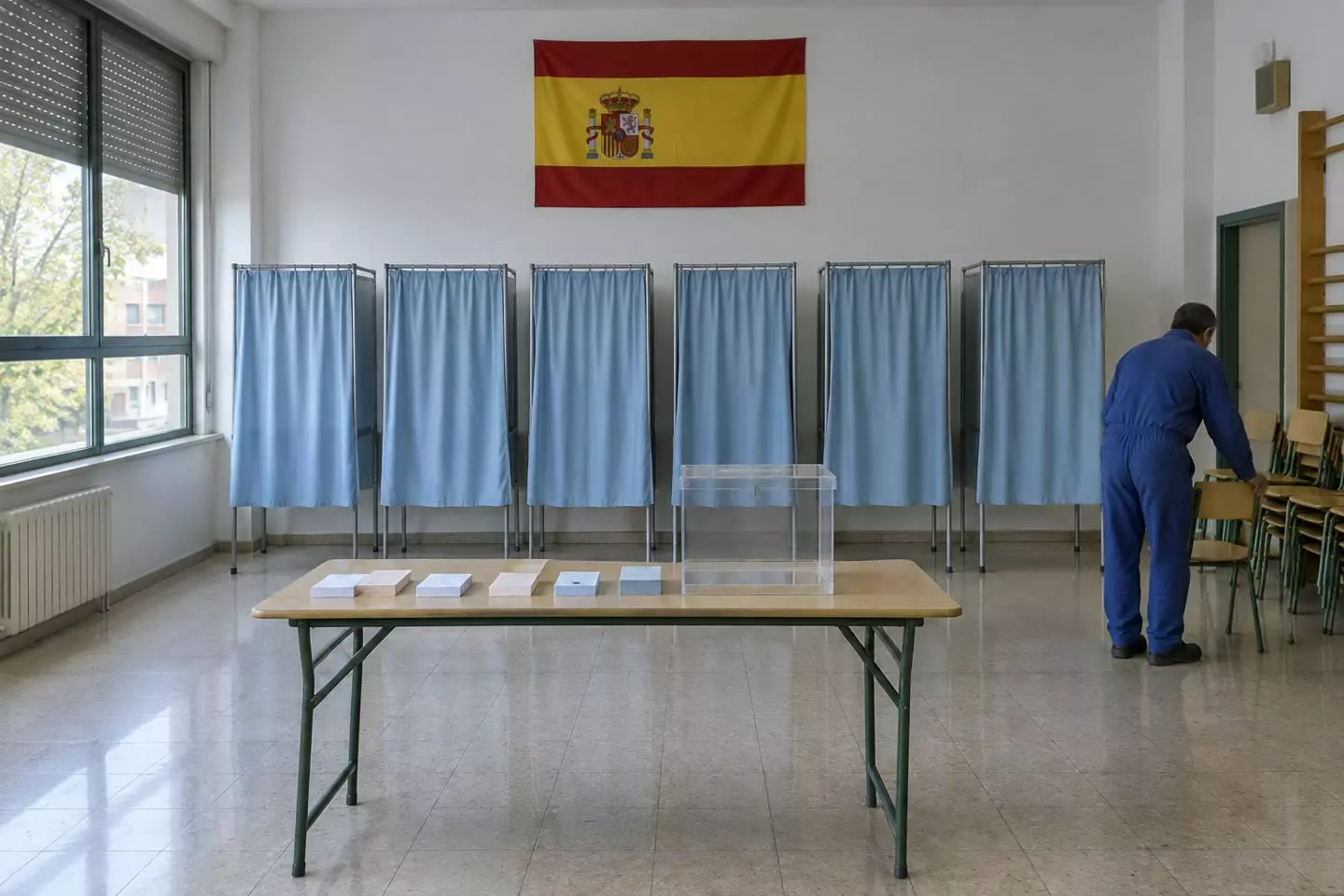 A polling station with blue-curtained booths and a transparent ballot box being prepared in a Madrid school hall in early autumn (illustration)