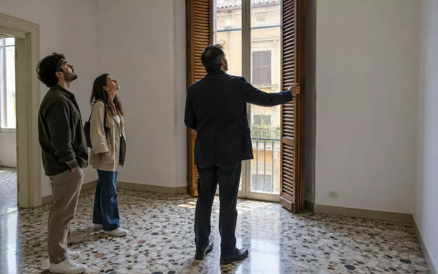 Ein Immobilienmakler in dunkler Jacke zeigt einem jungen Paar eine leere italienische Wohnung; alle drei sind von hinten aus mittlerer Entfernung zu sehen; hohe Fenster mit halb heruntergelassenen Holzläden und ein Terrazzoboden in sanftem Herbstlicht (Illustration).