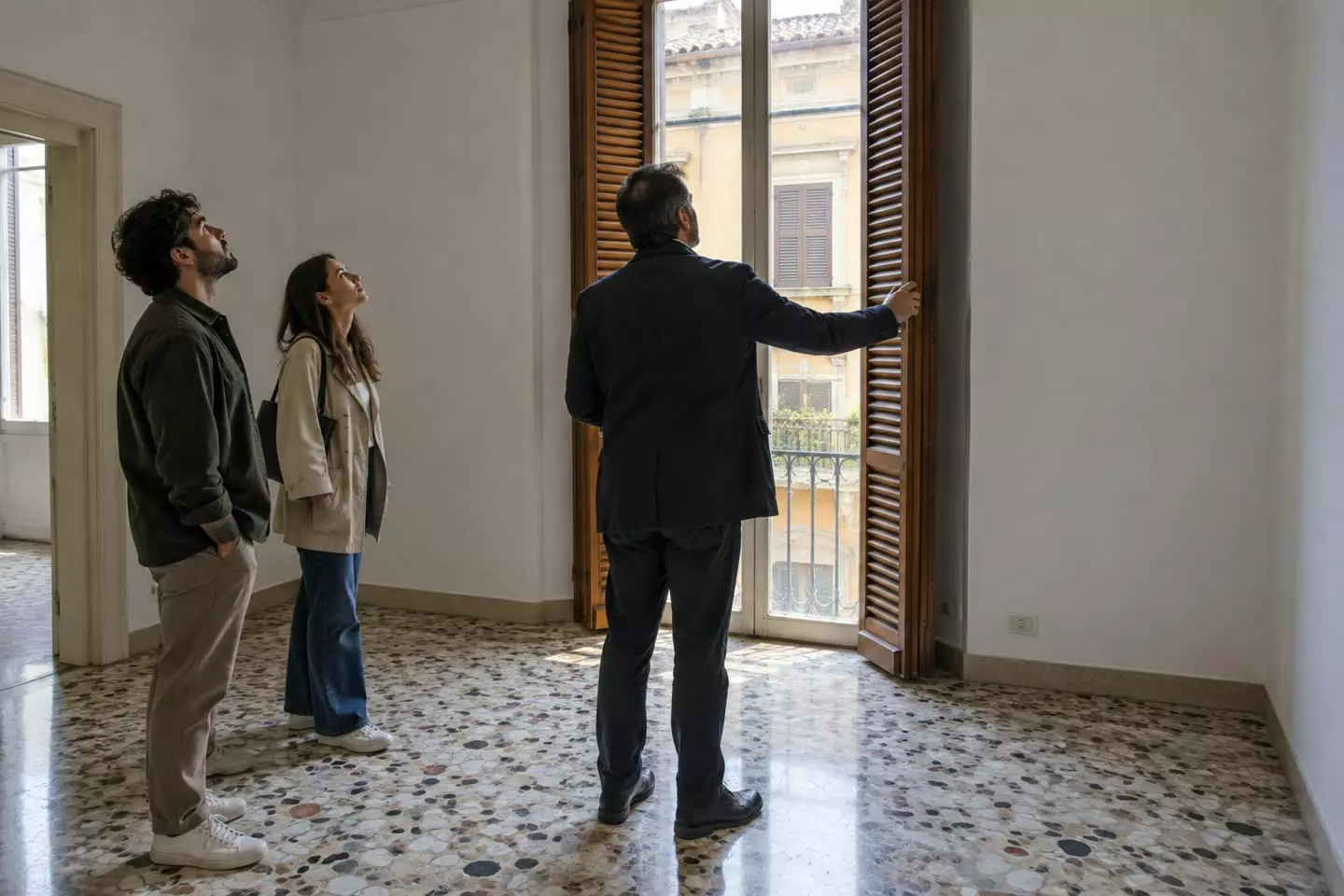 An estate agent in a dark jacket showing an empty Italian apartment to a young couple, all three seen from behind at mid-distance, tall windows with half-lowered wooden shutters and a terrazzo floor in soft autumn light (illustration)