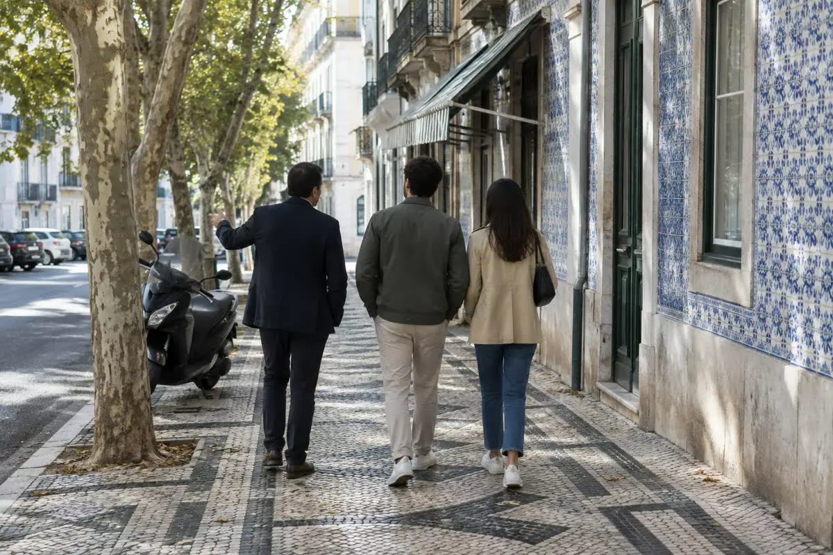 An estate agent in a dark jacket walking along a Lisbon pavement beside a young couple, all three seen from behind at mid-distance, an azulejo-tiled facade, a scooter at the kerb and a tiled Portuguese pavement in autumn light (illustration)