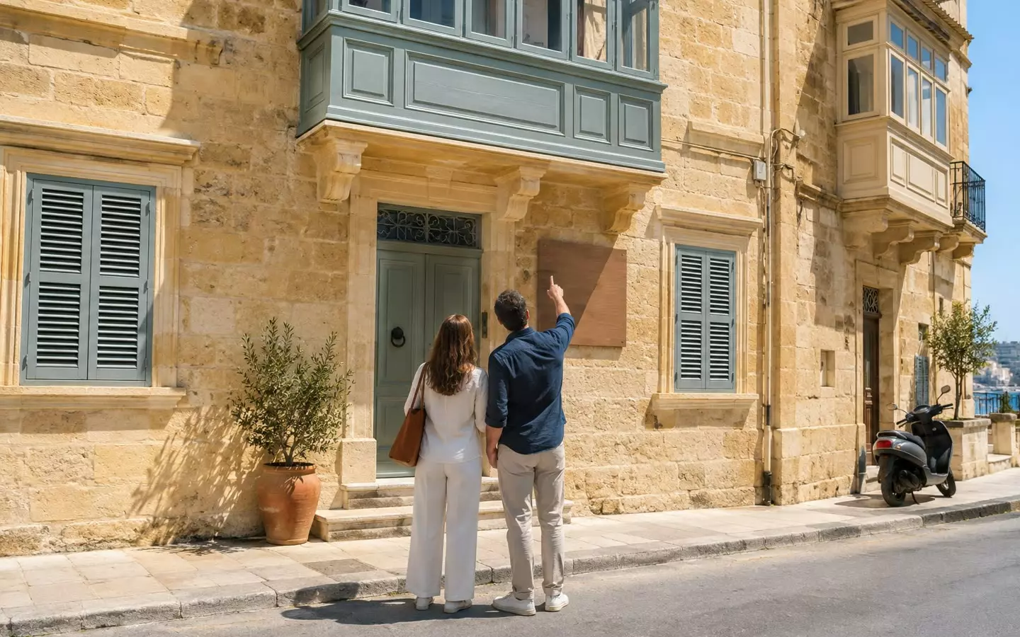 Ein Paar steht auf dem Bürgersteig vor einem maltesischen Stadthaus aus Kalkstein, an dessen Fassade ein Verkaufsschild angebracht ist. Das Haus ist von hinten aus mittlerer Entfernung zu sehen; darüber befindet sich ein bemalter, geschlossener Balkon und im Stockwerk darunter ein geöffneter Fensterladen (Abbildung).