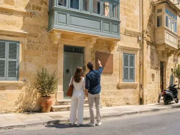 A couple standing on the pavement in front of a Maltese limestone townhouse with a sale board fixed to the facade, seen from behind at mid-distance, a painted enclosed balcony above them and a louvered shutter open on the floor below (illustration)