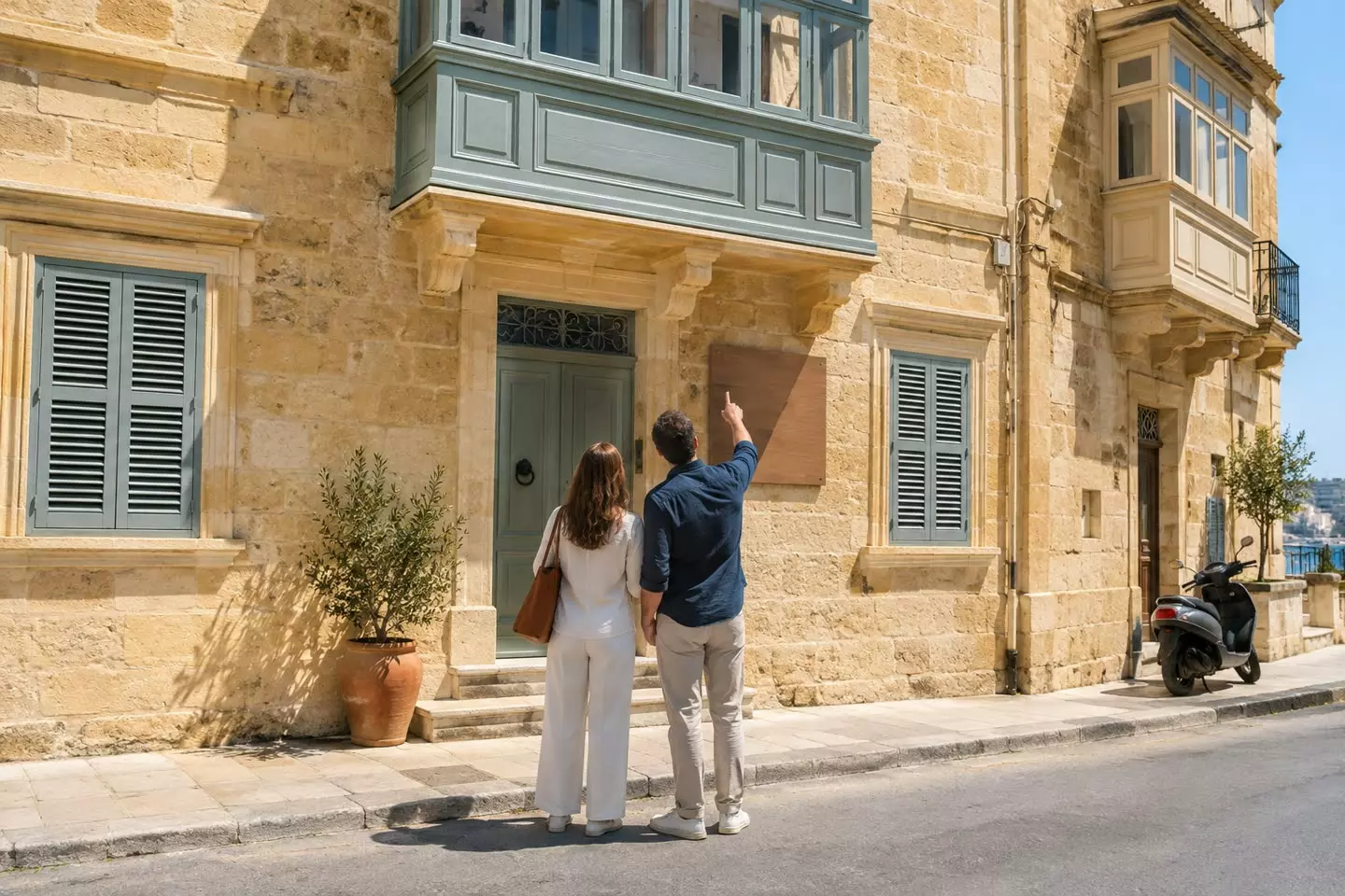 A couple standing on the pavement in front of a Maltese limestone townhouse with a sale board fixed to the facade, seen from behind at mid-distance, a painted enclosed balcony above them and a louvered shutter open on the floor below (illustration)