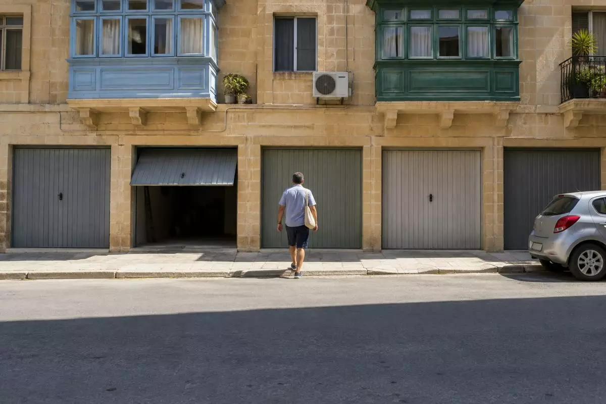 A row of metal garage doors set into the ground floor of a limestone apartment block in Malta with a man in a short-sleeved shirt seen from behind at mid-distance walking past them, balconies and an air-conditioning unit above (illustration)