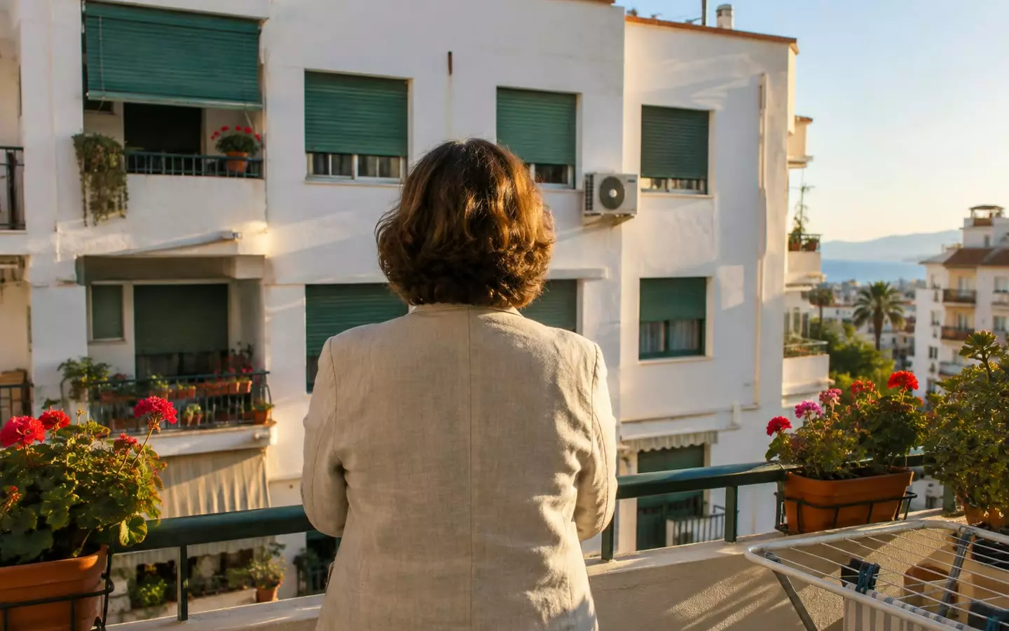 A woman standing on the balcony of an apartment in Malaga looking out over the neighbouring block, seen from behind at mid-distance, white rendered walls and green persianas in autumn light (illustration)