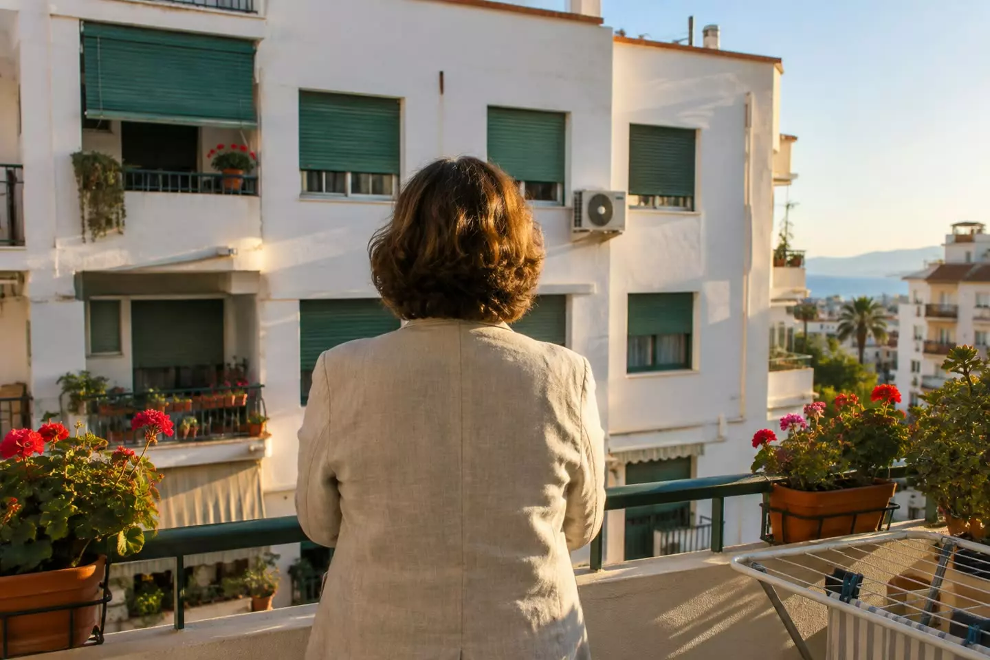 A woman standing on the balcony of an apartment in Malaga looking out over the neighbouring block, seen from behind at mid-distance, white rendered walls and green persianas in autumn light (illustration)