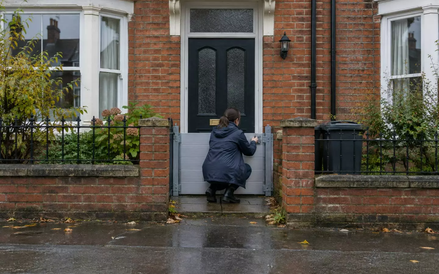 A woman in a raincoat standing with her back to the camera on the wet pavement outside an English red-brick terraced house, lifting a metal flood barrier into place across the front doorway at mid-distance, a low garden wall and a closed front door visible, overcast sky (illustration)