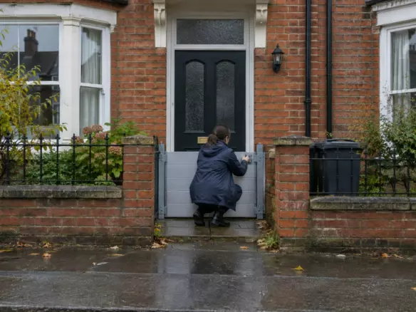 A woman in a raincoat standing with her back to the camera on the wet pavement outside an English red-brick terraced house, lifting a metal flood barrier into place across the front doorway at mid-distance, a low garden wall and a closed front door visible, overcast sky (illustration)