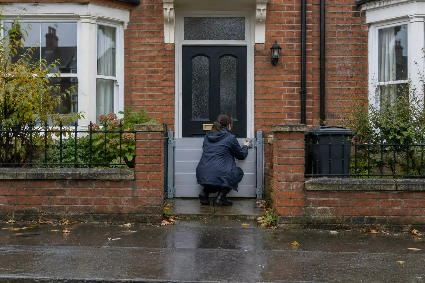 A woman in a raincoat standing with her back to the camera on the wet pavement outside an English red-brick terraced house, lifting a metal flood barrier into place across the front doorway at mid-distance, a low garden wall and a closed front door visible, overcast sky (illustration)