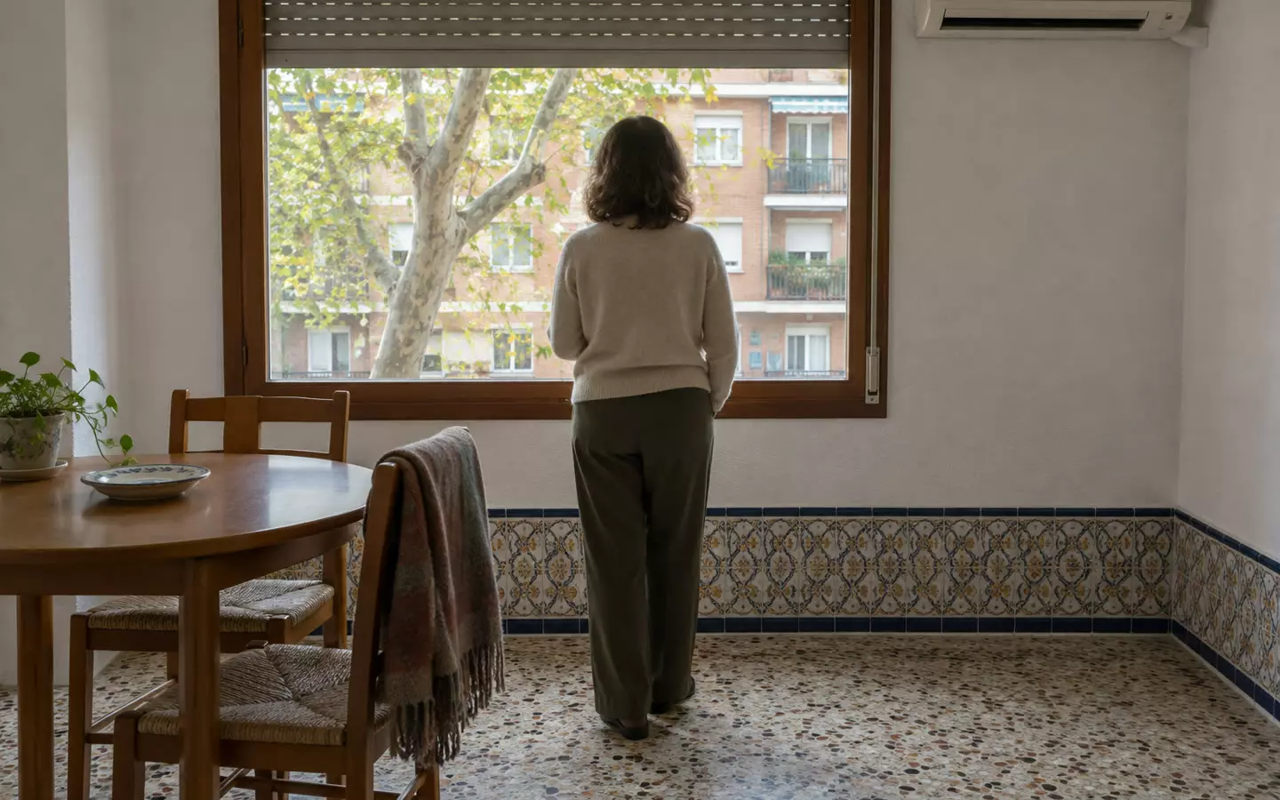 A woman in her fifties standing with her back to the camera at the window of a rented Madrid flat, one hand out of view, a half-raised persiana above her, a small round table with two chairs beside her and a split air-conditioning unit on the wall, a plane tree and the opposite block visible through the glass, mild October daylight (illustration)