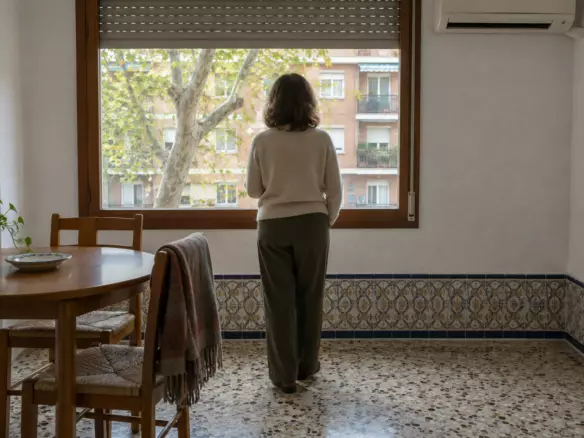 A woman in her fifties standing with her back to the camera at the window of a rented Madrid flat, one hand out of view, a half-raised persiana above her, a small round table with two chairs beside her and a split air-conditioning unit on the wall, a plane tree and the opposite block visible through the glass, mild October daylight (illustration)