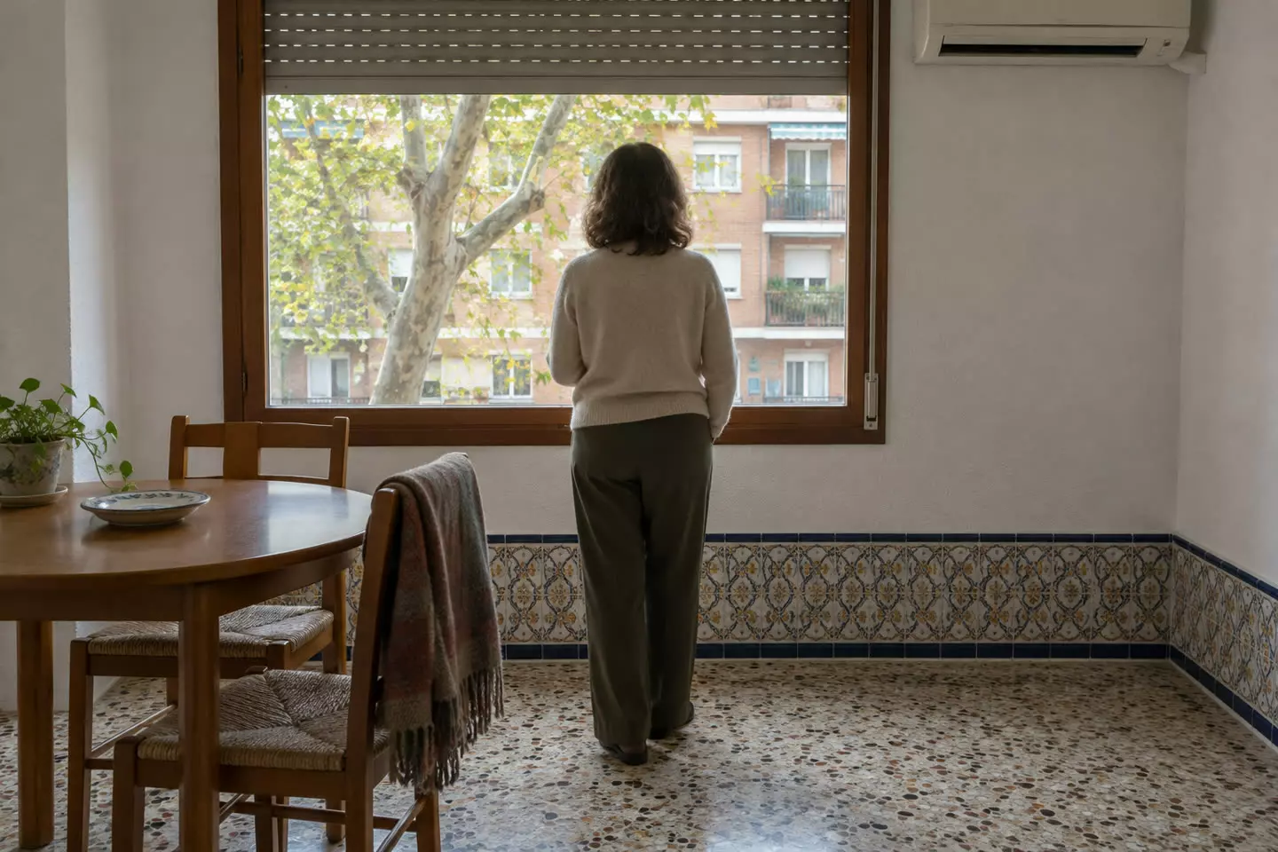 A woman in her fifties standing with her back to the camera at the window of a rented Madrid flat, one hand out of view, a half-raised persiana above her, a small round table with two chairs beside her and a split air-conditioning unit on the wall, a plane tree and the opposite block visible through the glass, mild October daylight (illustration)
