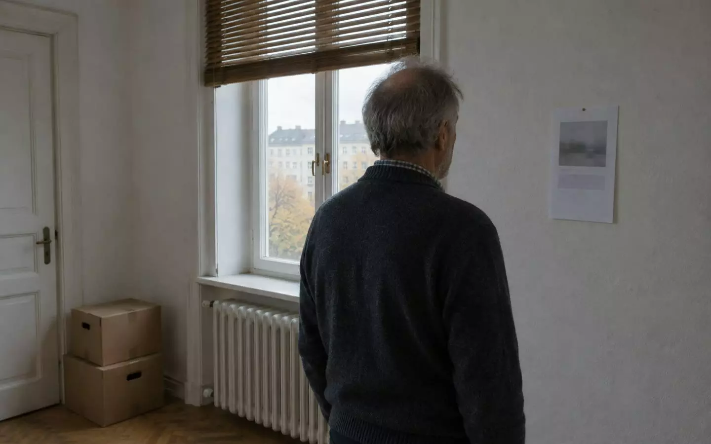 A man in his sixties seen three-quarters from behind at mid-distance in the living room of a rented Berlin Altbau flat, standing before a tall double window with a folded wooden roller blind and a white ribbed radiator below it, a printed notice pinned to the pale wall beside the window, soft grey autumn daylight (illustration)