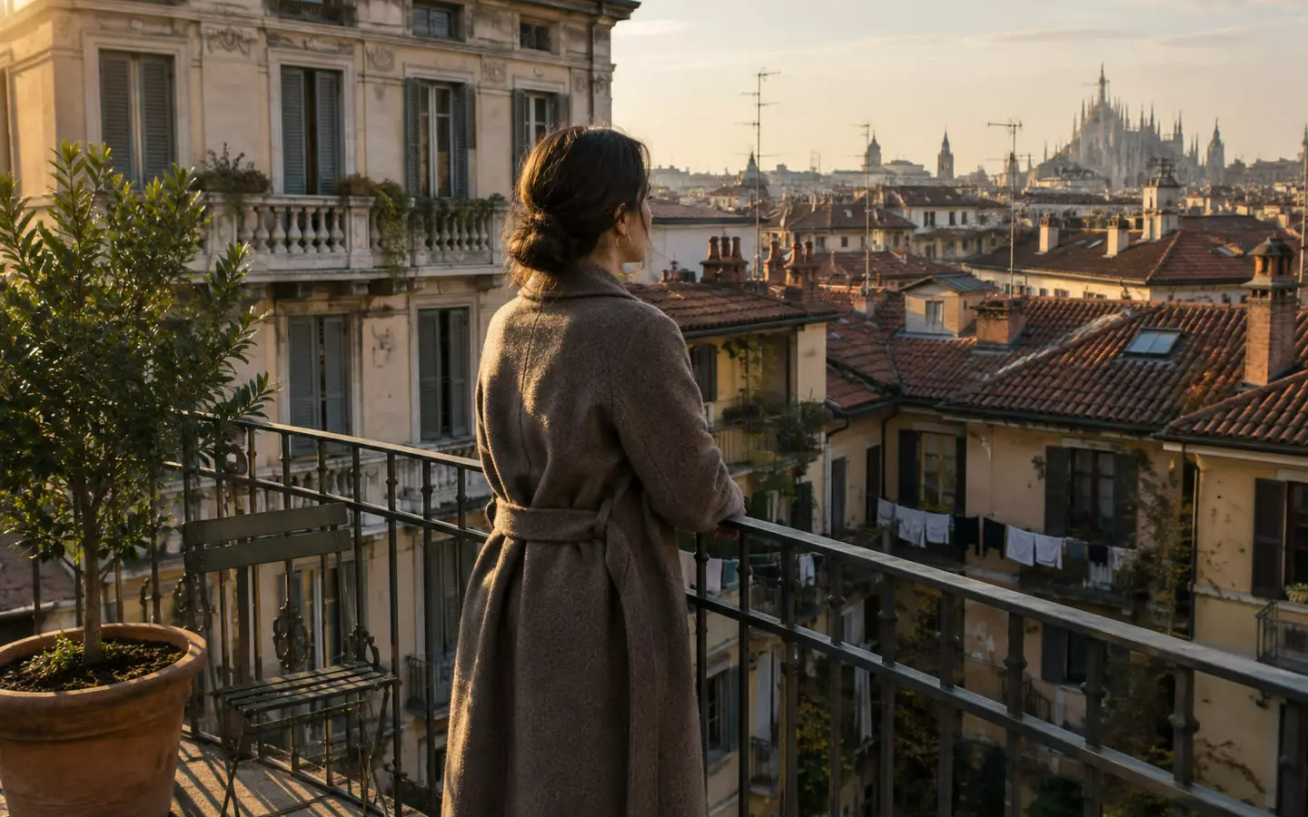 A woman in her thirties in a wool coat standing with her back three-quarters to the camera at the iron railing of a small rooftop terrace in Milan, looking out over a dense field of terracotta-tiled roofs, chimney pots, television antennas and a courtyard with washing lines below, a terracotta pot with a bay tree and a folded metal chair beside her, a 1930s Milanese apartment block with tall shuttered windows behind, warm low mid-October sun (illustration)