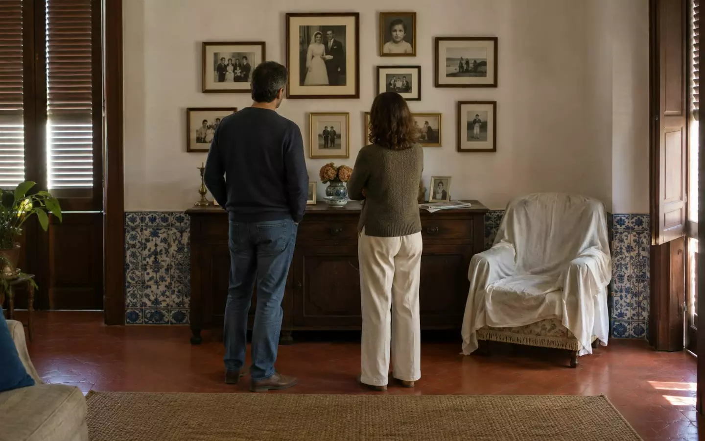 Two people in their forties standing with their backs to the camera at mid-distance in the living room of a Portuguese apartment, looking at a wall of framed family photographs above a dark wooden sideboard, an armchair under a white dust sheet beside them, a red terracotta floor and blue azulejo tiles along the skirting, half-closed wooden shutters with soft autumn light (illustration)