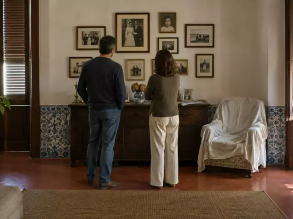 Two people in their forties standing with their backs to the camera at mid-distance in the living room of a Portuguese apartment, looking at a wall of framed family photographs above a dark wooden sideboard, an armchair under a white dust sheet beside them, a red terracotta floor and blue azulejo tiles along the skirting, half-closed wooden shutters with soft autumn light (illustration)