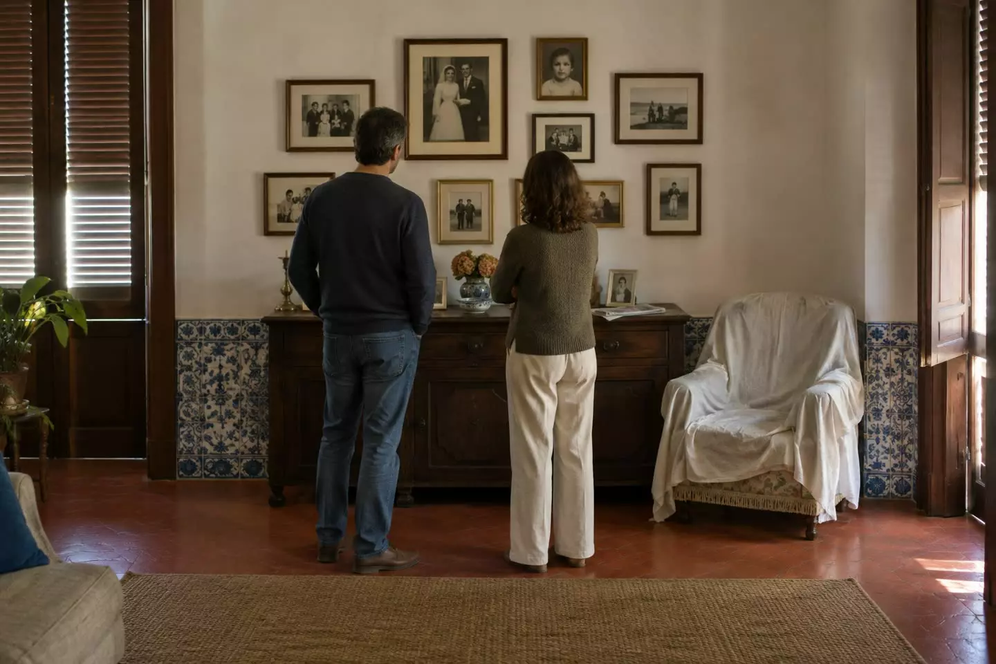 Two people in their forties standing with their backs to the camera at mid-distance in the living room of a Portuguese apartment, looking at a wall of framed family photographs above a dark wooden sideboard, an armchair under a white dust sheet beside them, a red terracotta floor and blue azulejo tiles along the skirting, half-closed wooden shutters with soft autumn light (illustration)