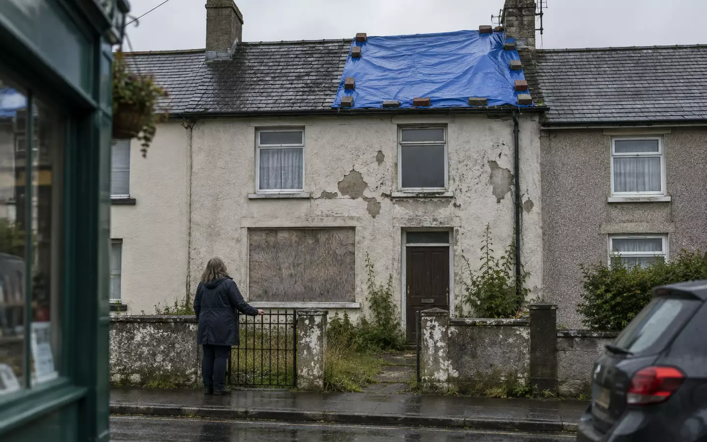 A woman in a dark raincoat stands at the garden gate of a two-storey Irish terraced house, seen from behind, its ground-floor window boarded with plywood and a blue tarpaulin weighted with bricks over part of the slate roof, peeling render, wet pavement and grey October light (illustration)