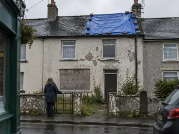 A woman in a dark raincoat stands at the garden gate of a two-storey Irish terraced house, seen from behind, its ground-floor window boarded with plywood and a blue tarpaulin weighted with bricks over part of the slate roof, peeling render, wet pavement and grey October light (illustration)