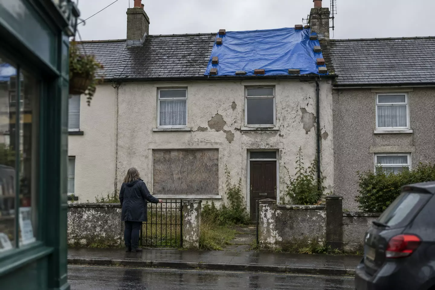 A woman in a dark raincoat stands at the garden gate of a two-storey Irish terraced house, seen from behind, its ground-floor window boarded with plywood and a blue tarpaulin weighted with bricks over part of the slate roof, peeling render, wet pavement and grey October light (illustration)