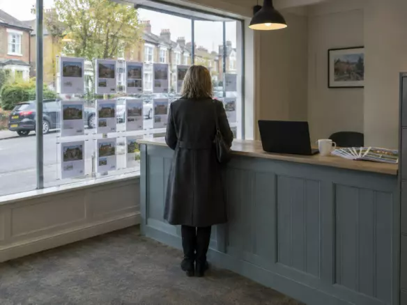 A woman in her fifties in a wool coat standing at a desk in a British high street estate agency, seen from behind at mid-distance, a window of property details in front of her, a laptop and a cup on the desk, grey October daylight through the glass (illustration)