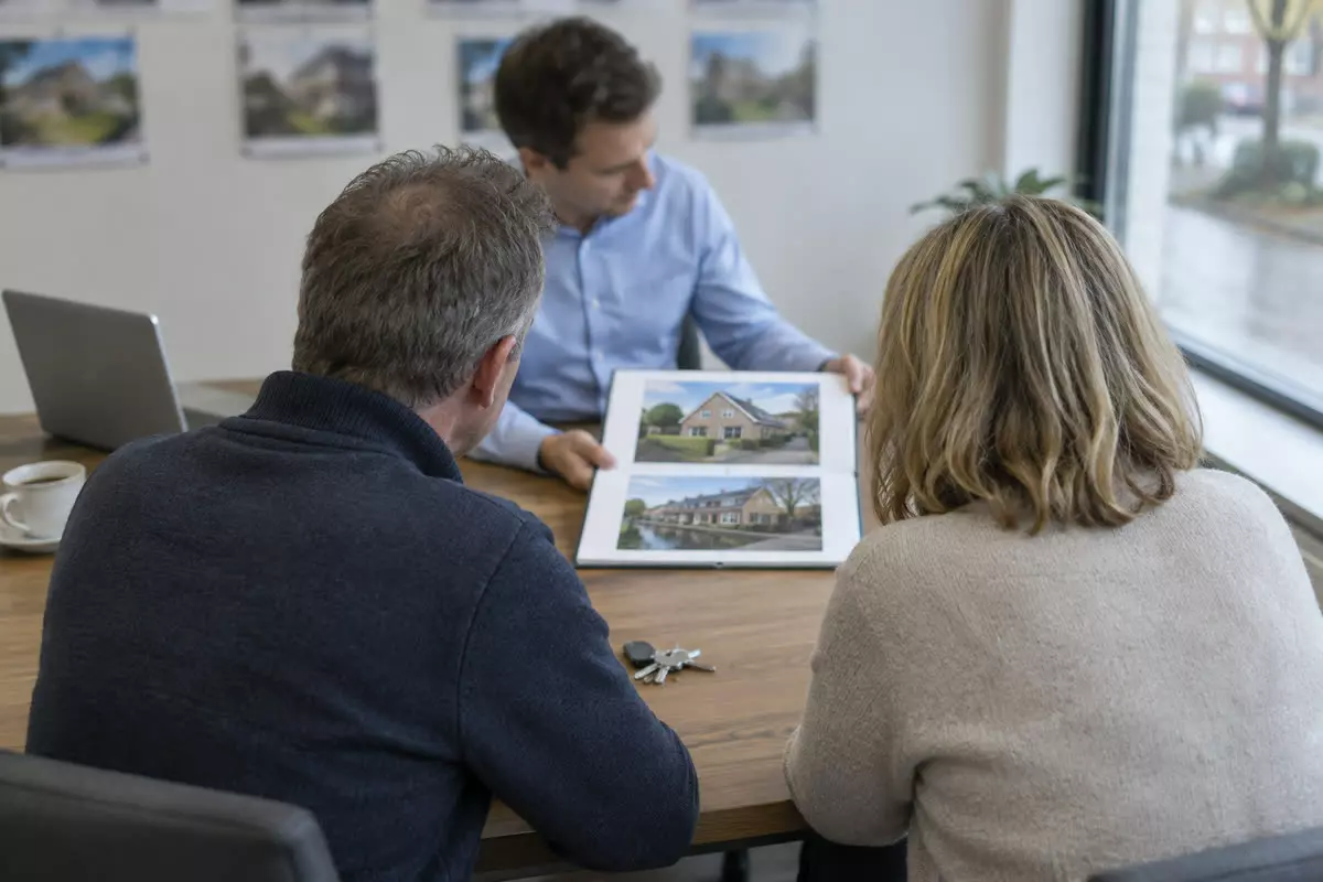 A couple in their fifties seated at a desk in a small Dutch estate agency office, seen from behind over their shoulders at mid-distance, an agent across the desk turning a folder of property photographs towards them, a laptop and a coffee cup on the desk, blurred house details pinned on the wall behind, pale autumn daylight through the shop window (illustration)