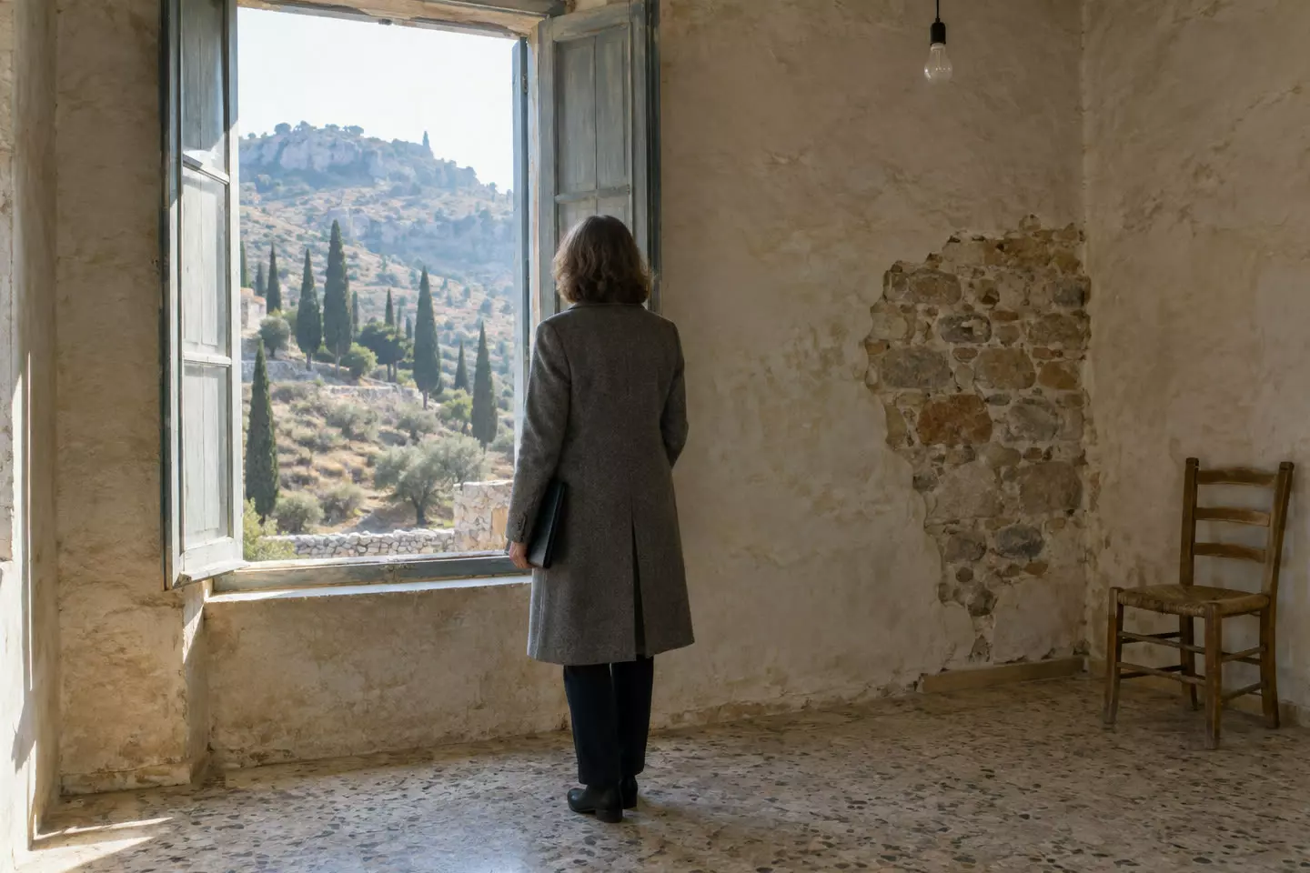 A woman in her fifties in a grey wool coat seen from behind at mid-distance inside an empty stone village house in the Peloponnese, a document folder under her arm, tall shutters open onto a dry hillside with cypresses, worn terrazzo floor and a bare pendant light (illustration)